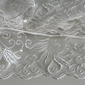Puede incluir: Borde de encaje blanco con un delicado patrón floral. El borde está doblado y apilado, mostrando los intrincados detalles del diseño.