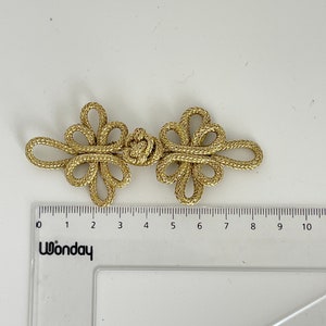 May include: A pair of gold-colored Chinese knot closures, also known as frog closures, are shown against a white background. The knots are approximately 3 inches long and are made of a braided cord.