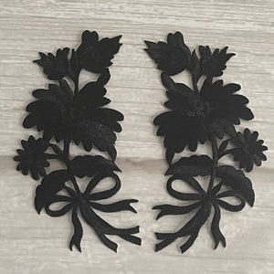 Peut inclure: Deux appliques en tissu noir en forme de bouquets floraux avec des nœuds. Les appliques sont découpées et présentent un design délicat et complexe.