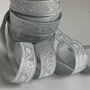 May include: A roll of silver ribbon featuring a floral pattern. The ribbon is a light grey with a metallic finish. The floral design is white and intricate. The ribbon is stacked, with some unrolled ribbon visible in the foreground.
