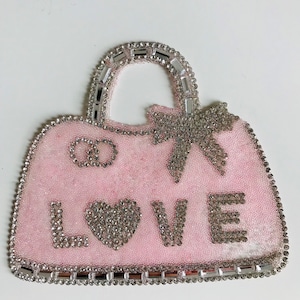 Peut inclure: Un sac à main en tissu rose avec un ornement en strass et le mot "LOVE" en strass. Le sac à main a une poignée et un nœud.