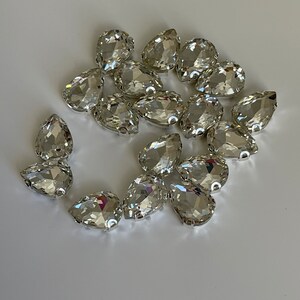 May include: A group of clear crystal teardrop-shaped rhinestones, scattered on a white surface.