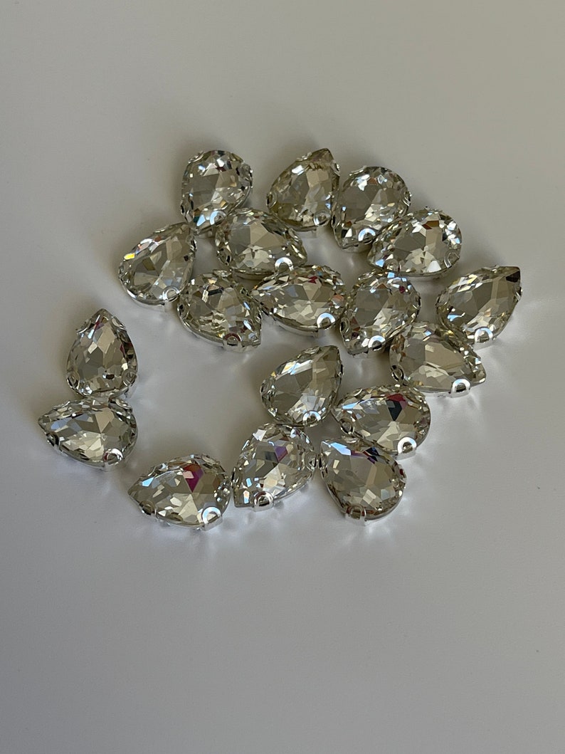 May include: A group of clear crystal teardrop-shaped rhinestones, scattered on a white surface.