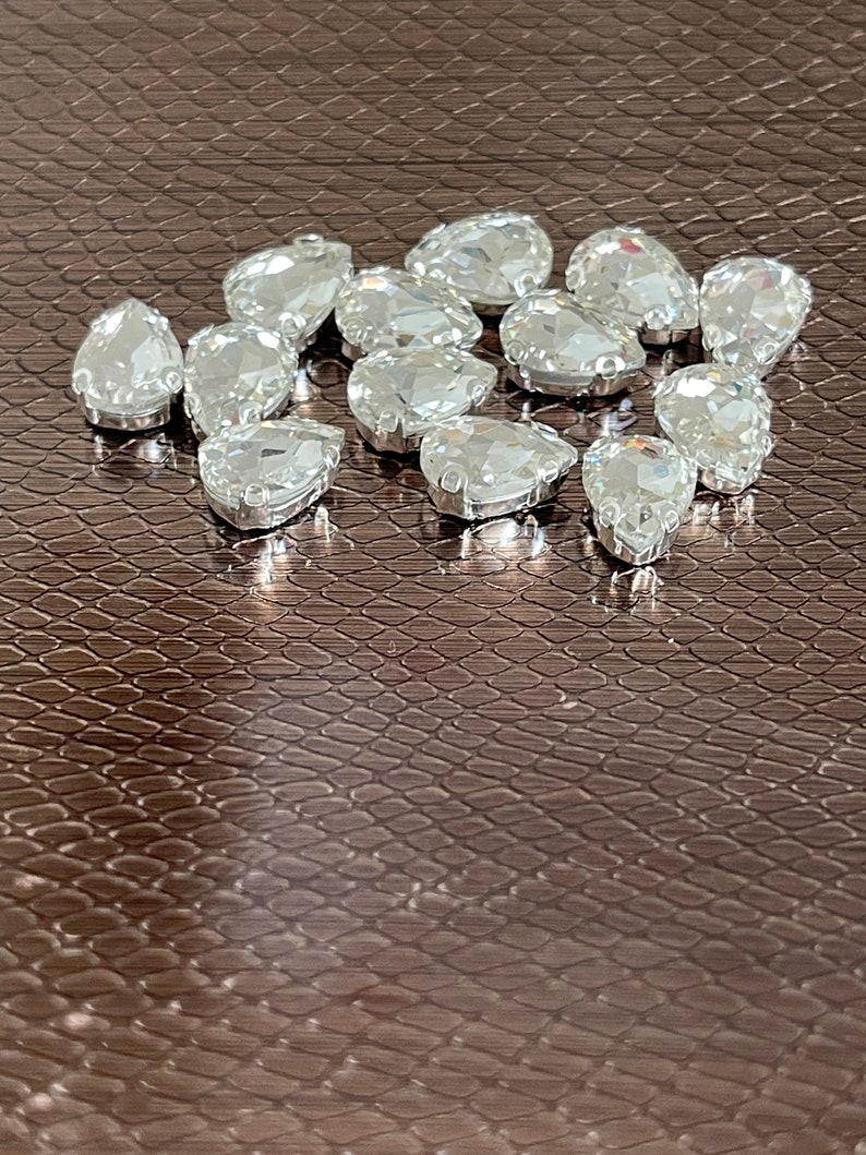 May include: A group of clear teardrop-shaped rhinestones on a brown textured surface.