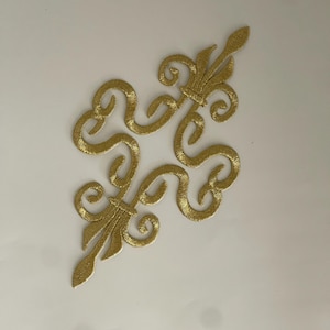 Peut inclure: Deux écussons brodés dorés en forme de fleur de lys. Chaque écusson présente un motif de lys stylisé avec des volutes complexes et une base pointue. Les écussons sont placés sur un fond blanc.