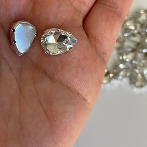 May include: Two clear glass pear-shaped rhinestones with silver settings. One rhinestone is in its setting, the other is loose.