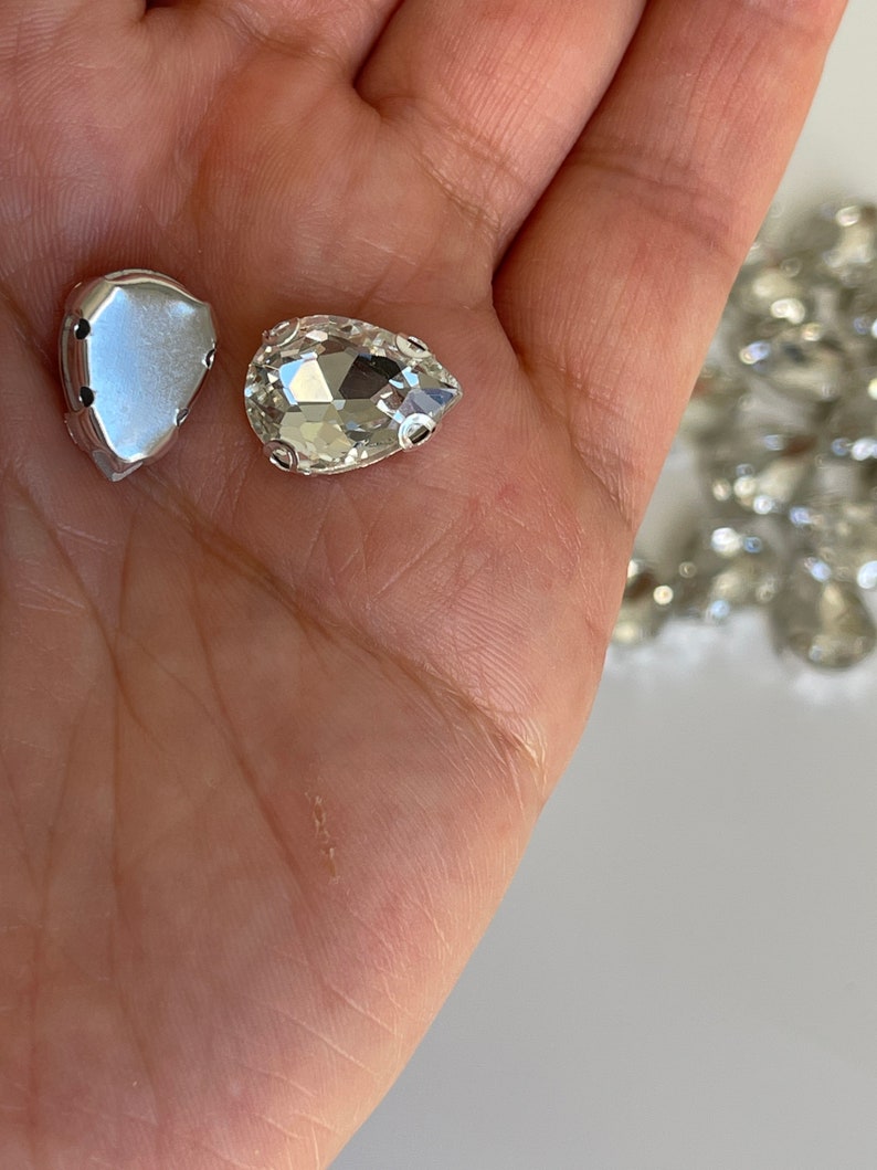May include: Two clear glass pear-shaped rhinestones with silver settings. One rhinestone is in its setting, the other is loose.