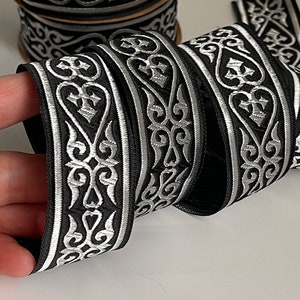 Peut inclure: Ruban noir et argent avec un motif répété de croix et de volutes stylisées. Le ruban mesure environ 2,5 cm de large.