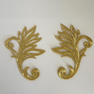 Peut inclure: Deux appliques en forme de feuilles brodées en fil doré. Chaque applique présente un motif détaillé avec une tige centrale et des segments en forme de feuilles, ainsi qu'un élément décoratif enroulé. Sur fond blanc.