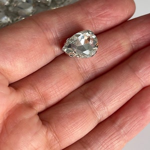 May include: A clear teardrop-shaped crystal rhinestone with a silver setting.