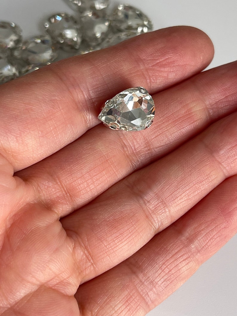 May include: A clear teardrop-shaped crystal rhinestone with a silver setting.