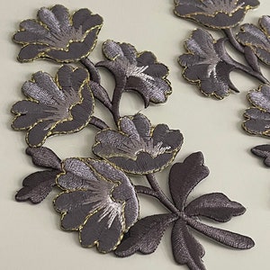 Peut inclure: Deux ensembles d'appliques en tissu brodées représentant des fleurs grises avec des accents dorés. Les fleurs sont disposées en grappe avec des feuilles.