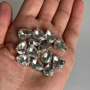 May include: A close-up of a hand holding a pile of clear teardrop-shaped rhinestones. The rhinestones have a silver backing and are perfect for adding sparkle to crafts and jewelry.
