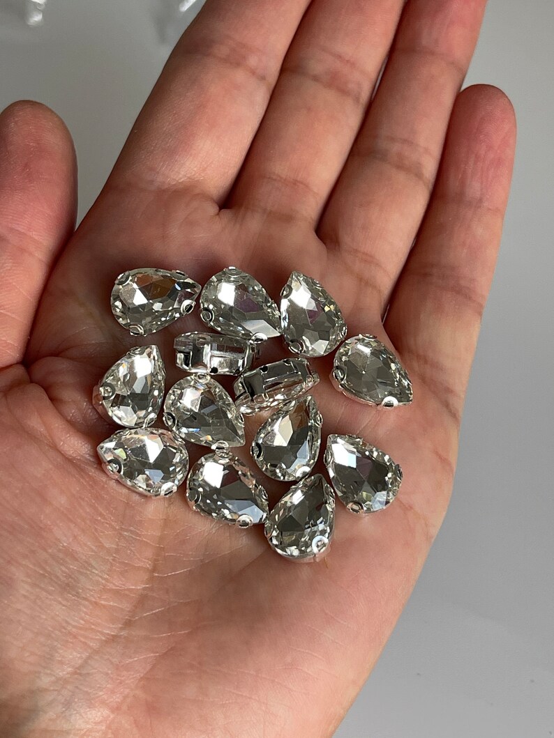 May include: A close-up of a hand holding a pile of clear teardrop-shaped rhinestones. The rhinestones have a silver backing and are perfect for adding sparkle to crafts and jewelry.