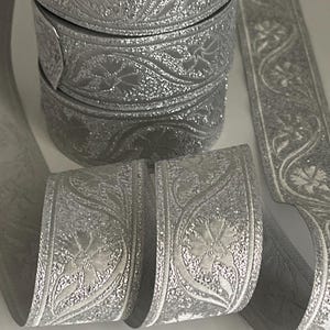 Könnte beinhalten: Silbernes Zierband mit floralem Design. Das Band ist zu mehreren Spulen gerollt und abgerollt, wodurch das aufwendige geprägte Muster sichtbar wird. Das Band hat eine glitzernde, strukturierte Oberfläche und verleiht Eleganz.