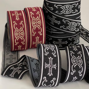 May include: Close-up of several spools of decorative ribbon. The ribbons feature repeating patterns of crosses and ornate designs in various colours, including burgundy, black, and grey. The ribbon is likely used for crafting or embellishment.