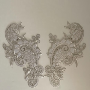 May include: Two silver lace appliques in a paisley design with floral accents. The appliques are ideal for adding a touch of elegance to any project.