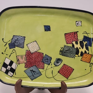 Può includere: Un vassoio rettangolare in ceramica con sfondo verde chiaro e bordo nero. Il vassoio è decorato con quadrati colorati a motivi e linee nere ondulate. Il vassoio è sostenuto da un supporto in legno marrone.
