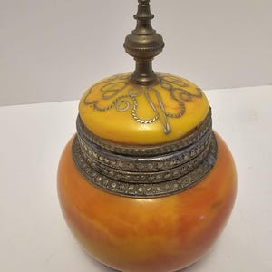 May include: A round, orange and yellow glass jar with a brass lid and a decorative silver band around the top. The lid has a small, pointed brass finial.