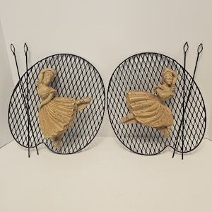 Op de afbeelding: Twee zwarte metalen wandsculpturen met een rasterpatroon. Elke sculptuur heeft een beige ballerina-figuur in een dansende pose.