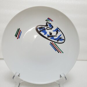 May include: White ceramic bowl with a blue, black and gold abstract design. The design features a stylised paint palette with black and blue lines and a gold outline. The bowl has a red, green and black stripe design on the side.