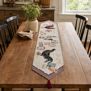 May include: A rectangular table runner featuring a cream-colored fabric with printed illustrations of roosters, vintage postage stamps, and cursive script. The runner has a red and white checkered border and a small red tassel at the pointed end.