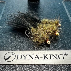May include: Three fly fishing lures with gold beads and black and brown feathers. The lures are on a black surface with a silver label that reads "DYNA-KING".