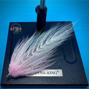 May include: A handmade fly fishing lure with a pink and white feather design. The lure is mounted on a black base with the text "DYNA-KING" and the logo "FW Fly on the Water Bespoke Flies". The background is a solid blue.