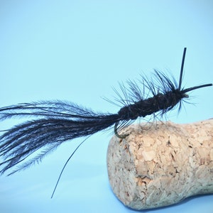 May include: A black fly fishing lure with a long tail of feathers, tied to a cork.