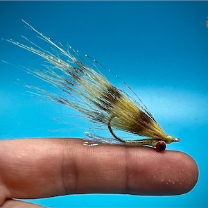May include: A close-up of a fly fishing lure with a brown and white body and a red bead. The lure is tied on a hook and is being held by a finger.