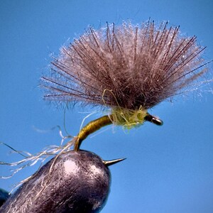 May include: A close-up of a fishing fly with a dark hook and a fluffy, brown head. The fly has a yellow body and white, wispy tail fibers. The background is a solid, light blue.