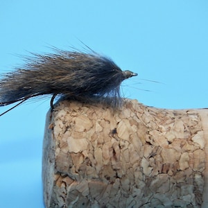 May include: A dark gray and brown fly fishing lure with a brown feather body and a silver hook. The lure is sitting on a cork.