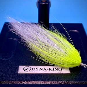 May include: A handmade fly fishing lure with a bright yellow and white feathered body. The lure is attached to a silver hook and is mounted on a black base with the Dyna-King logo.