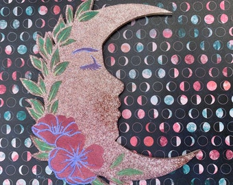 Pode incluir: Uma lua de madeira em forma de crescente com acabamento brilhante, pintada com um rosto estilizado e detalhes florais. A lua é decorada com flores vermelhas e roxas e folhas verdes.