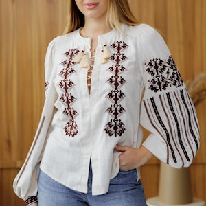 Pode incluir: Blusa de linho branco com bordados castanhos intrincados na frente, mangas e punhos. A blusa apresenta um decote em V com detalhes de borlas e mangas compridas bufantes. A modelo está usando jeans azuis.
