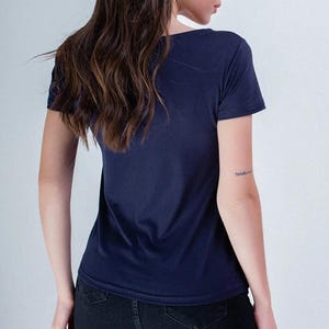 May include: A navy blue t-shirt with a crew neck. The model is wearing black jeans. The person has long, wavy brown hair. A tattoo is visible on the left arm. The background is a plain white wall.