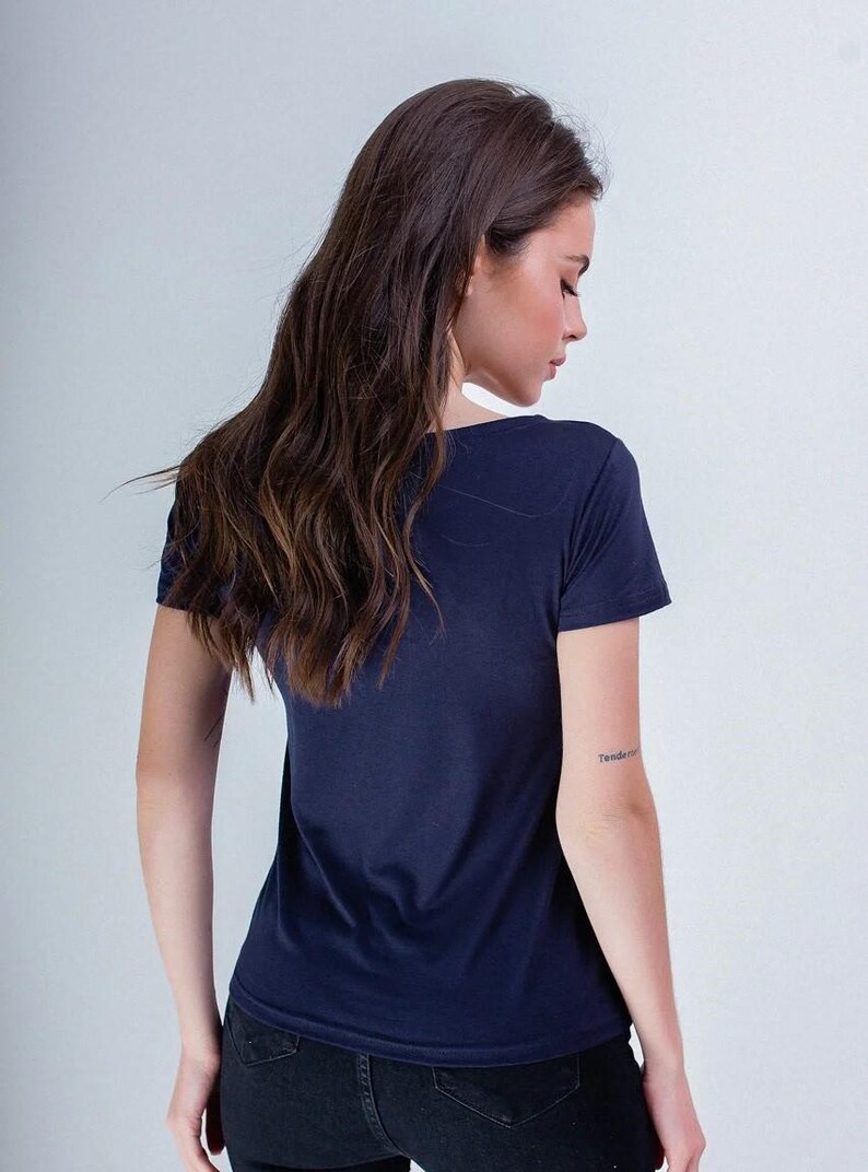 May include: A navy blue t-shirt with a crew neck. The model is wearing black jeans. The person has long, wavy brown hair. A tattoo is visible on the left arm. The background is a plain white wall.