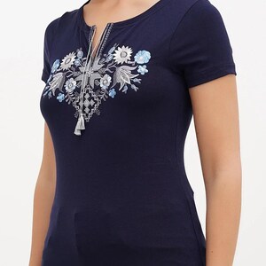 May include: Navy blue short-sleeve t-shirt with a scoop neckline and a decorative floral embroidery. The embroidery features white, light blue, and gray flowers and leaves, with a tassel detail at the neckline.