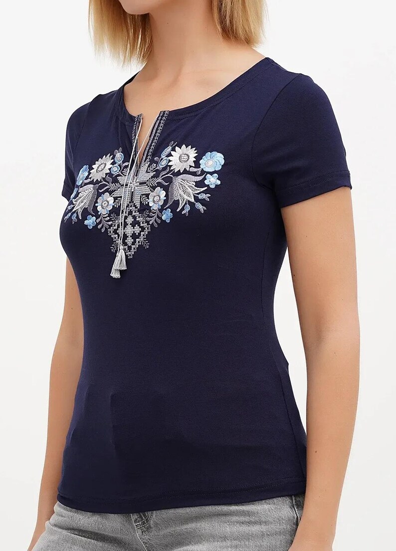 May include: Navy blue short-sleeve t-shirt with a scoop neckline and a decorative floral embroidery. The embroidery features white, light blue, and gray flowers and leaves, with a tassel detail at the neckline.
