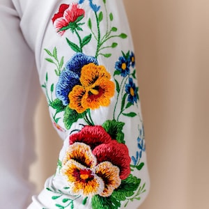 Close-up detail of a white 3/4 length sleeve showcasing beautiful machine embroidery of colorful pansies and wildflowers in Spanish style.