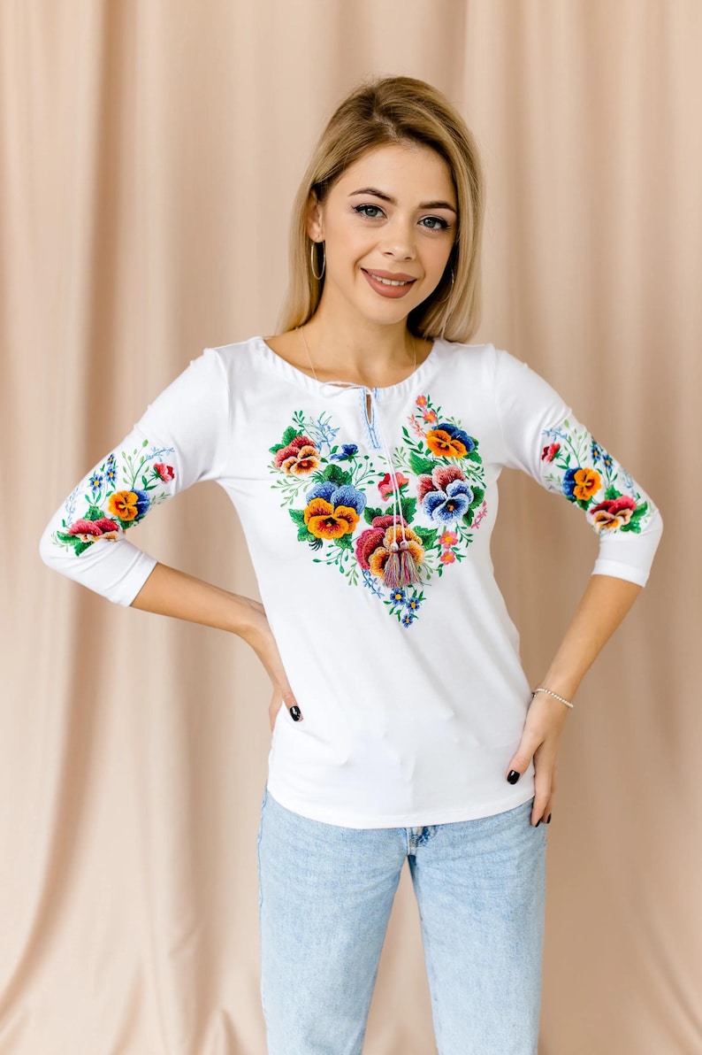 Front view of a white embroidered hippie tee with a keyhole neckline, colorful tassels, and a rich botanical pansy design.