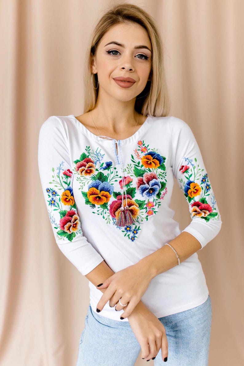 Studio portrait of a blonde woman wearing a bright white ethnic peasant shirt featuring vibrant purple, yellow, and red floral embroidery.