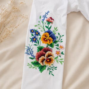 Flat lay close-up of a white viscose sleeve displaying bright multicolored floral satin-stitch embroidery against a neutral background.