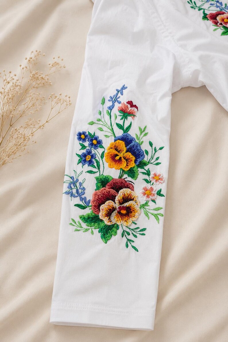 Flat lay close-up of a white viscose sleeve displaying bright multicolored floral satin-stitch embroidery against a neutral background.