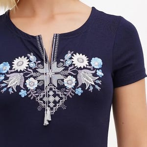 May include: Navy blue short-sleeve shirt with floral embroidery in white, light blue, and gray. The embroidered design is centered on the chest and features a keyhole neckline with tassel details. The shirt has a round neckline.