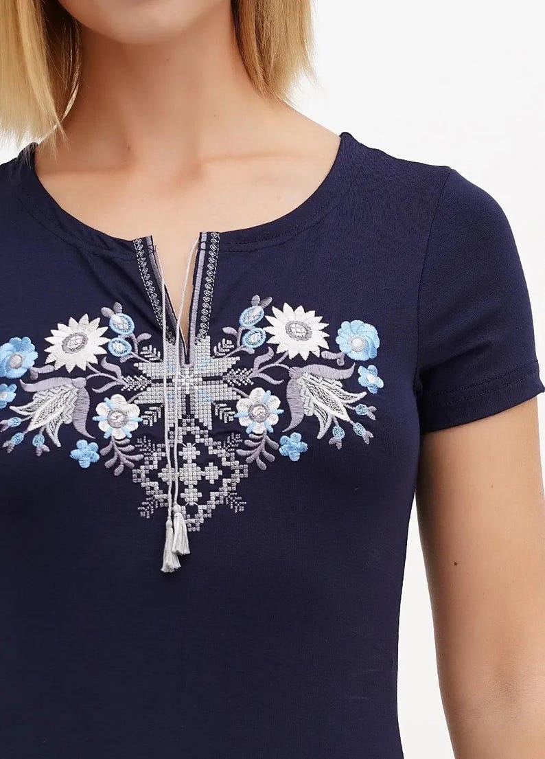 May include: Navy blue short-sleeve shirt with floral embroidery in white, light blue, and gray. The embroidered design is centered on the chest and features a keyhole neckline with tassel details. The shirt has a round neckline.
