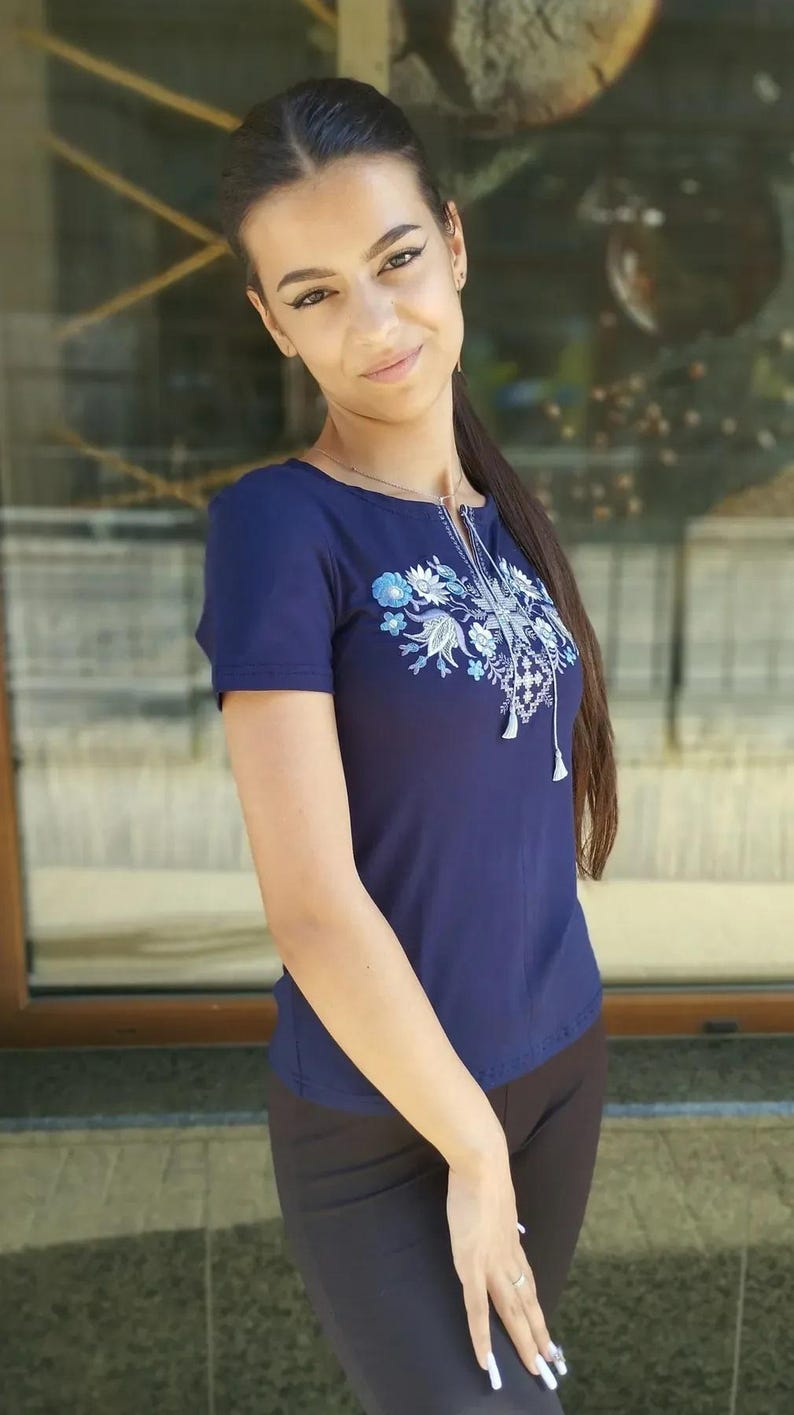 May include: A navy blue short-sleeved top with floral embroidery in white and light blue. The top has a round neckline and a decorative tassel. The person is wearing black pants.