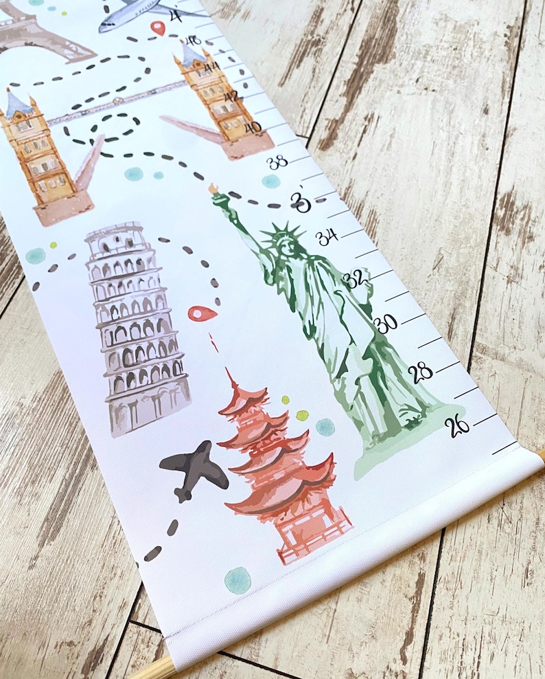 Puede incluir: Un medidor de altura de lienzo blanco con ilustraciones en acuarela de monumentos mundiales, incluyendo la Torre Eiffel, el Big Ben, la Torre de Pisa, la Estatua de la Libertad y una pagoda, con una ruta de viaje punteada y marcas en pulgadas.