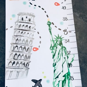 Puede incluir: Un medidor de altura blanco con ilustraciones en acuarela de la Torre de Pisa, la Estatua de la Libertad y un puente. Un camino de viaje en l&iacute;nea negra discontinua conecta los monumentos, junto a un peque&ntilde;o avi&oacute;n y marcas numeradas en cent&iacute;metros.