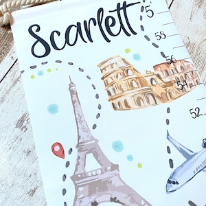 Puede incluir: Un medidor de altura de tela blanca para colgar con una cuerda, que presenta el nombre Scarlett en letra negra, ilustraciones de la Torre Eiffel y el Coliseo, un avi&oacute;n azul y una escala vertical marcada en cm del 52 al 60.